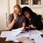 Casal sentado à mesa, analisando documentos e consultando um smartphone enquanto organiza as finanças domésticas.