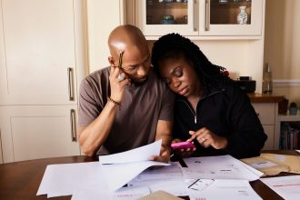 Casal sentado à mesa, analisando documentos e consultando um smartphone enquanto organiza as finanças domésticas.