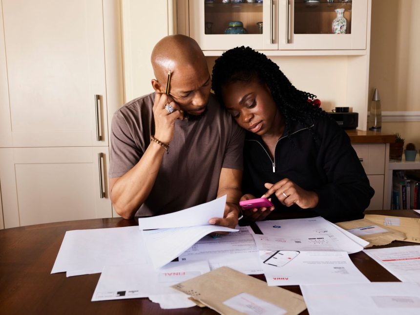 Casal sentado à mesa, analisando documentos e consultando um smartphone enquanto organiza as finanças domésticas.